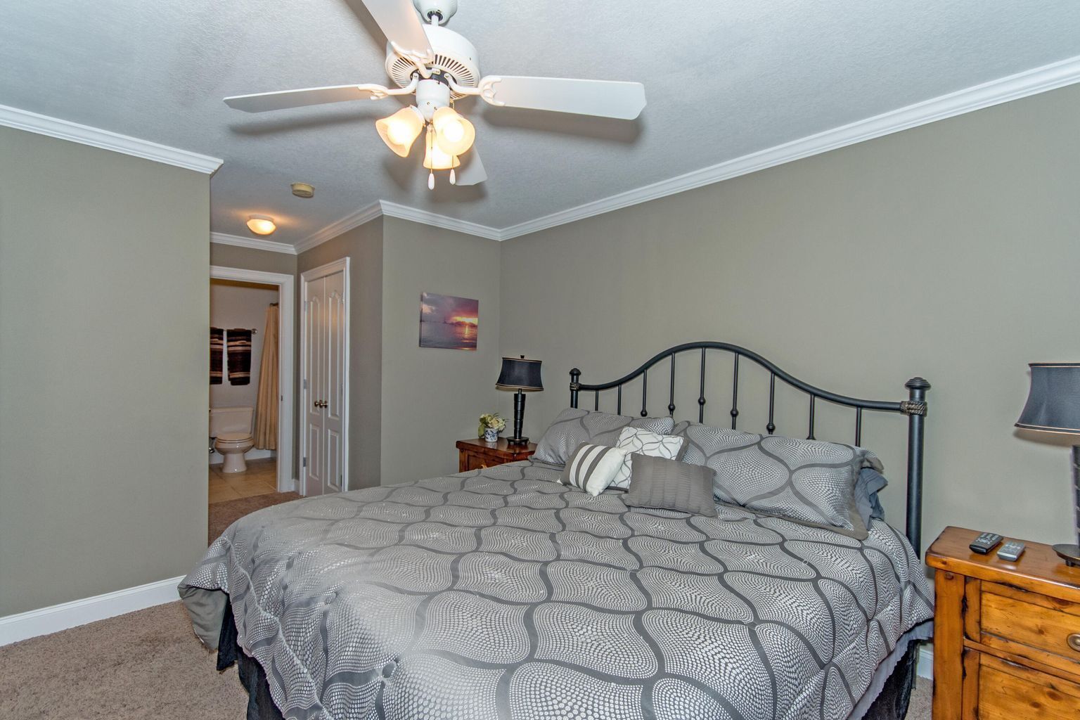 A bedroom with a bed, a nightstand, a lamp, a ceiling fan, and a door leading to a bathroom.