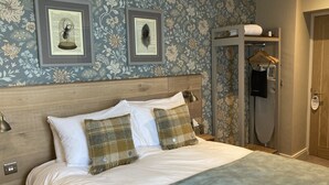 A neatly made bed with white linens and plaid pillows, a wooden headboard, and a wall with floral wallpaper. - Old Swan (Skipton)