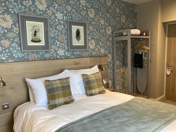 A neatly made bed with white linens and plaid pillows, a wooden headboard, and a wall with floral wallpaper.