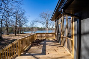 Une terrasse en bois, dotée d’une rambarde, mène à un bâtiment aux grandes fenêtres, qui offre une vue sur un lac et des arbres.