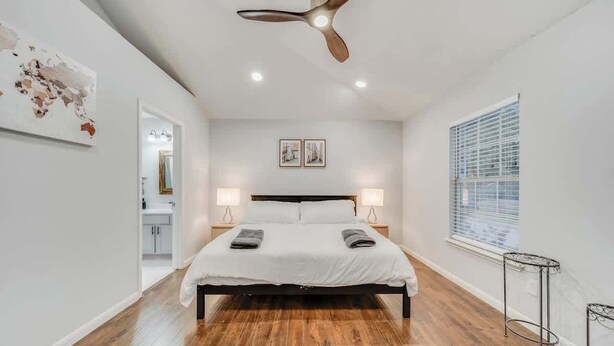 Une chambre avec un plancher en bois, un lit recouvert de draps blancs, deux lampes de chevet, un ventilateur de plafond et une fenêtre avec des stores.