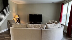Un salon comprenant un canapé d’angle aux lignes courbes, un téléviseur à écran plat, une lampe et un escalier.