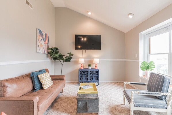 A living room with a brown leather couch, a striped armchair, a small wooden table with a tray, a blue cabinet with two lamps, and a flat-screen TV on the wall. - Spacious ranch 65” 4K tv deck, grill, fire pit fenced yd , office,  baby gear (Cape May Court House)