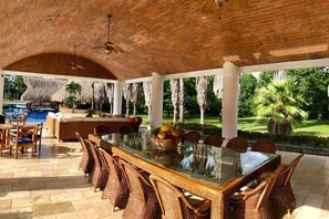 Uma área de refeições ao ar livre com uma mesa de vidro, cadeiras de vime e uma piscina ao fundo.