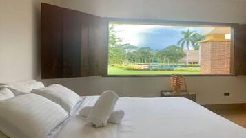 Quarto de hotel com uma cama grande, roupa de cama branca, vista para uma piscina e vegetação, e uma estrutura de tijolos visível pela janela.