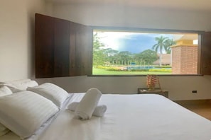 Quarto de hotel com uma cama grande, roupa de cama branca, vista para uma piscina e vegetação, e uma estrutura de tijolos visível pela janela.
