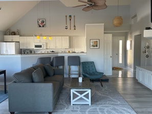 A modern living room with a grey sofa, a teal armchair, and a coffee table. A kitchen with white cabinets and a microwave is visible in the background.