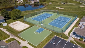 An aerial view of a tennis court complex with two courts, a parking area, and a surrounding landscape including a pond and residential buildings.