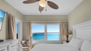 Un dormitorio con una cama grande, ventilador de techo, vista al océano y una ventana con cortinas.