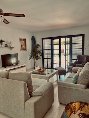 Ein Wohnzimmer mit Fernseher, Sofas, einem Couchtisch und einem großen Fenster mit Jalousien.