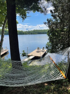 Eine Hängematze zwischen Bäumen, mit Blick auf einen See mit einem Steg und Stühlen.