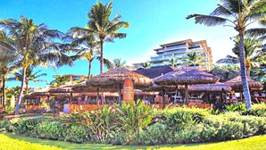Un resort con estructuras de techo de paja, palmeras y un edificio de varios pisos al fondo.