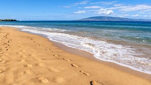 A beach with footprints leading to the water, a clear sky, and distant mountains.