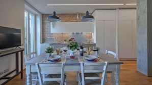 A dining area with a long table set for four, two pendant lights above, and a kitchen in the background.