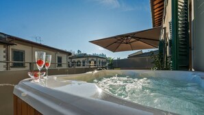 Un bain à remous avec deux verres de vin rouge, posés sur une terrasse en bois, un parasol et une vue sur les bâtiments en arrière-plan.