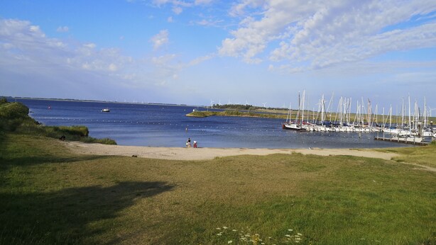 Un port de plaisance avec des bateaux amarrés, une plage de sable et des éoliennes visibles au loin.
