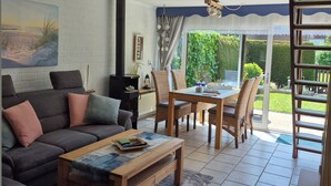 Un salon comprenant un canapé, une table basse et une table à manger avec des chaises. On y trouve également une cheminée, une fenêtre avec des rideaux et une vue sur la verdure à l’extérieur.