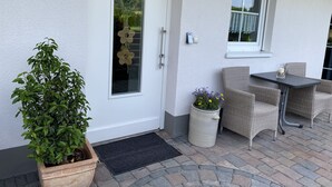 Pared exterior blanca con una puerta, una planta en maceta, una mesa pequeña con sillas y una ventana que ofrece vistas a zonas verdes.