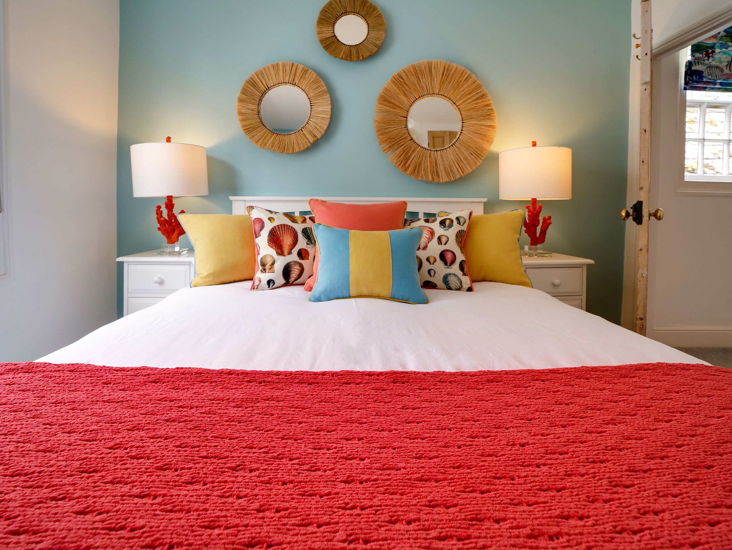 Un lit recouvert d’une couverture rouge à texture, d’une couette blanche et d’oreillers colorés ornés d’un motif de coquillage. Deux miroirs encadrés d’un motif tissé sont fixés au mur, et deux lampes de chevet sont posées sur des tables de chevet blanches.