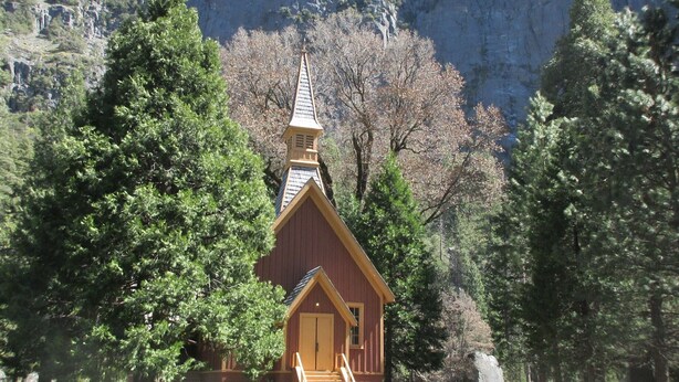 Una pequeña capilla de madera con techo empinado, rodeada de árboles frondosos y un paisaje montañoso.