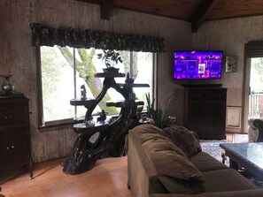 Un salon doté d’un plancher en bois, d’un canapé, d’une télévision et d’une grande fenêtre donnant sur la forêt.