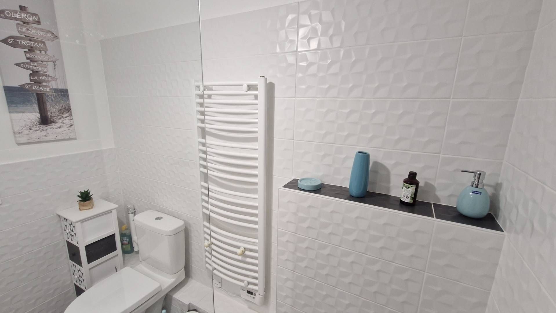 Ein Badezimmer mit weiß gefliester Wand, einem weißen Heizkörper, einer Toilette, einem Waschbecken und einem Ablagebereich für Pflegeartikel.