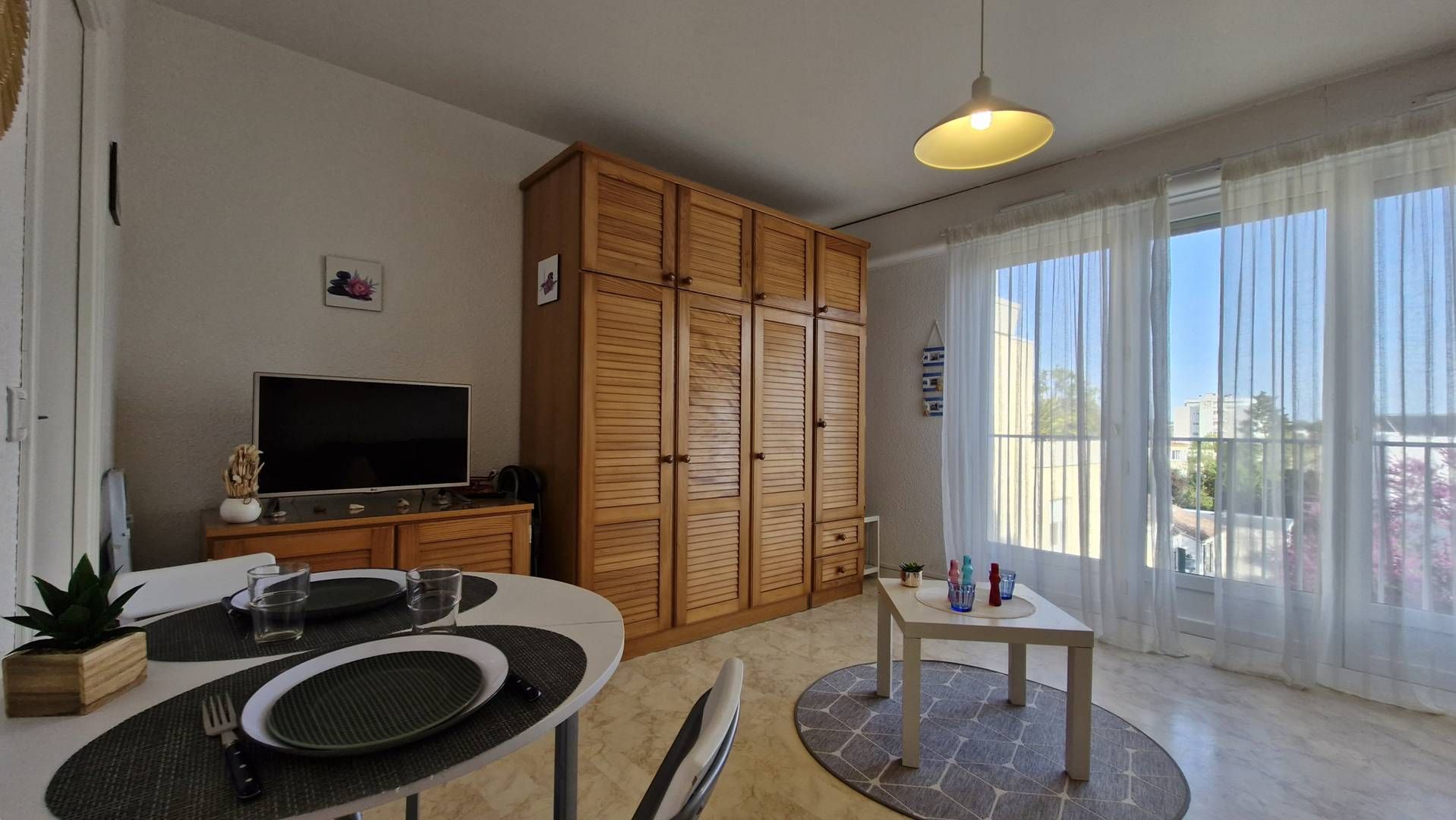 Een moderne woonkamer met een eethoek, een houten garderobe, een televisie en grote ramen met gordijnen.