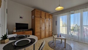 Ein modernes Wohnzimmer mit Essbereich, einem Holzschrank, Fernseher und großen Fenstern mit Vorhängen.