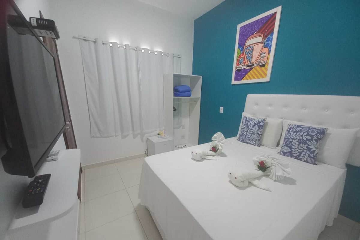 A hotel room with a bed, a TV, a curtain, a shelf, and a painting on the wall.