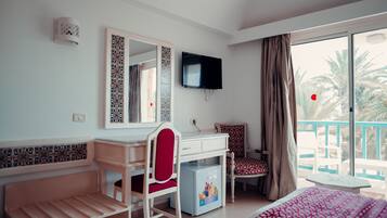 Ein Hotelzimmer mit Schreibtisch, Stuhl, Bett, Fernseher und einem Balkon mit Blick auf Palmen.