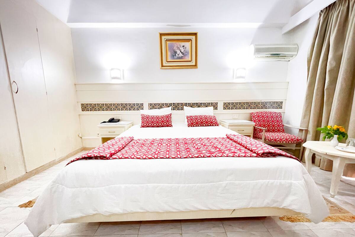 Une chambre d’hôtel avec un grand lit, une table de chevet, une chaise, une petite table ornée d’un vase de fleurs et un climatiseur fixé au mur.