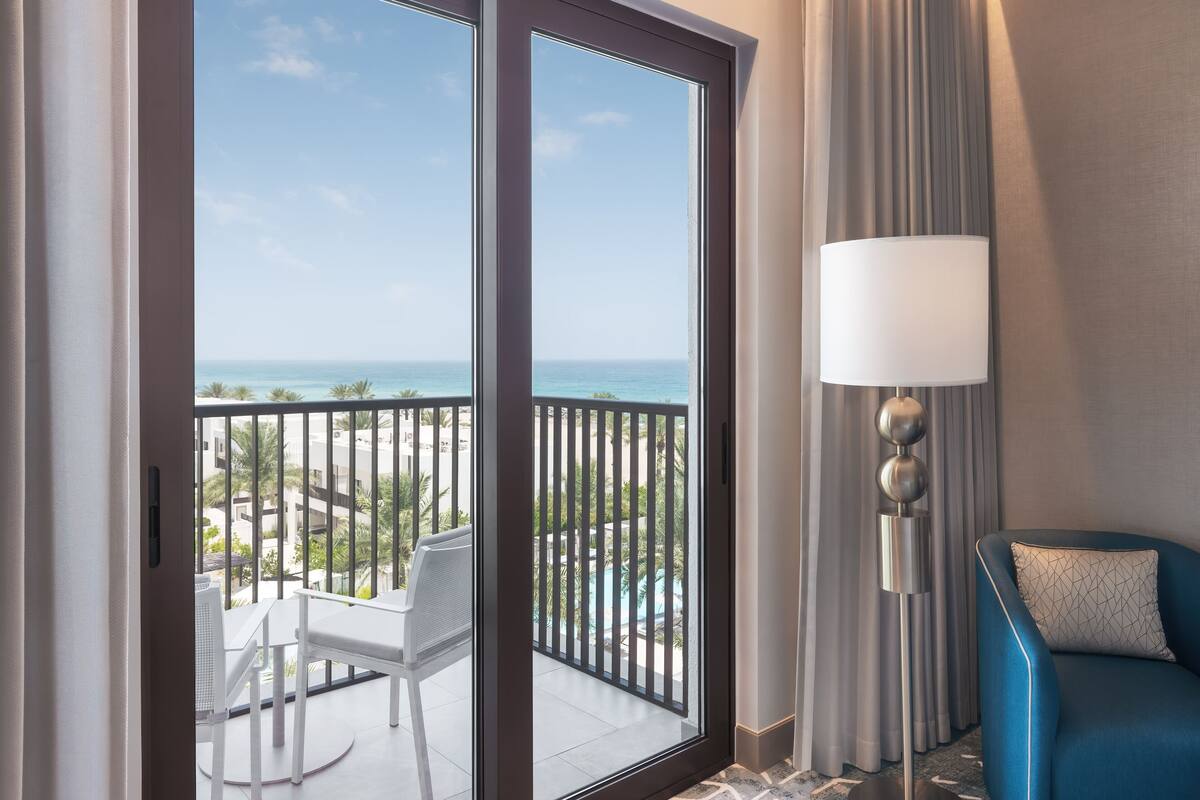 Un balcon avec vue sur l’océan, une chaise blanche, un canapé bleu et une lampe sur pied.