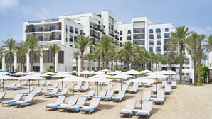 Un hôtel en bord de mer, avec plusieurs étages, une plage de sable dotée de transats et de parasols, et des palmiers.