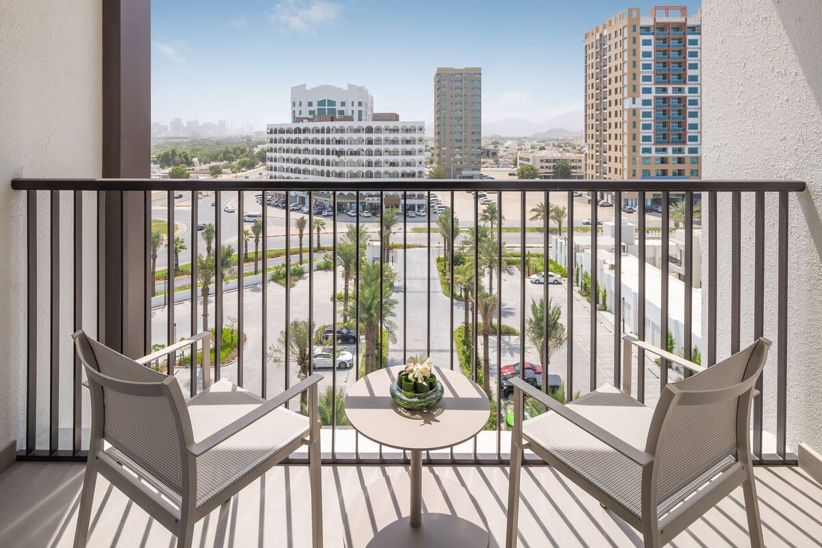 Un balcon avec deux chaises et une petite table, d’où l’on peut admirer un paysage urbain avec ses bâtiments et sa route.