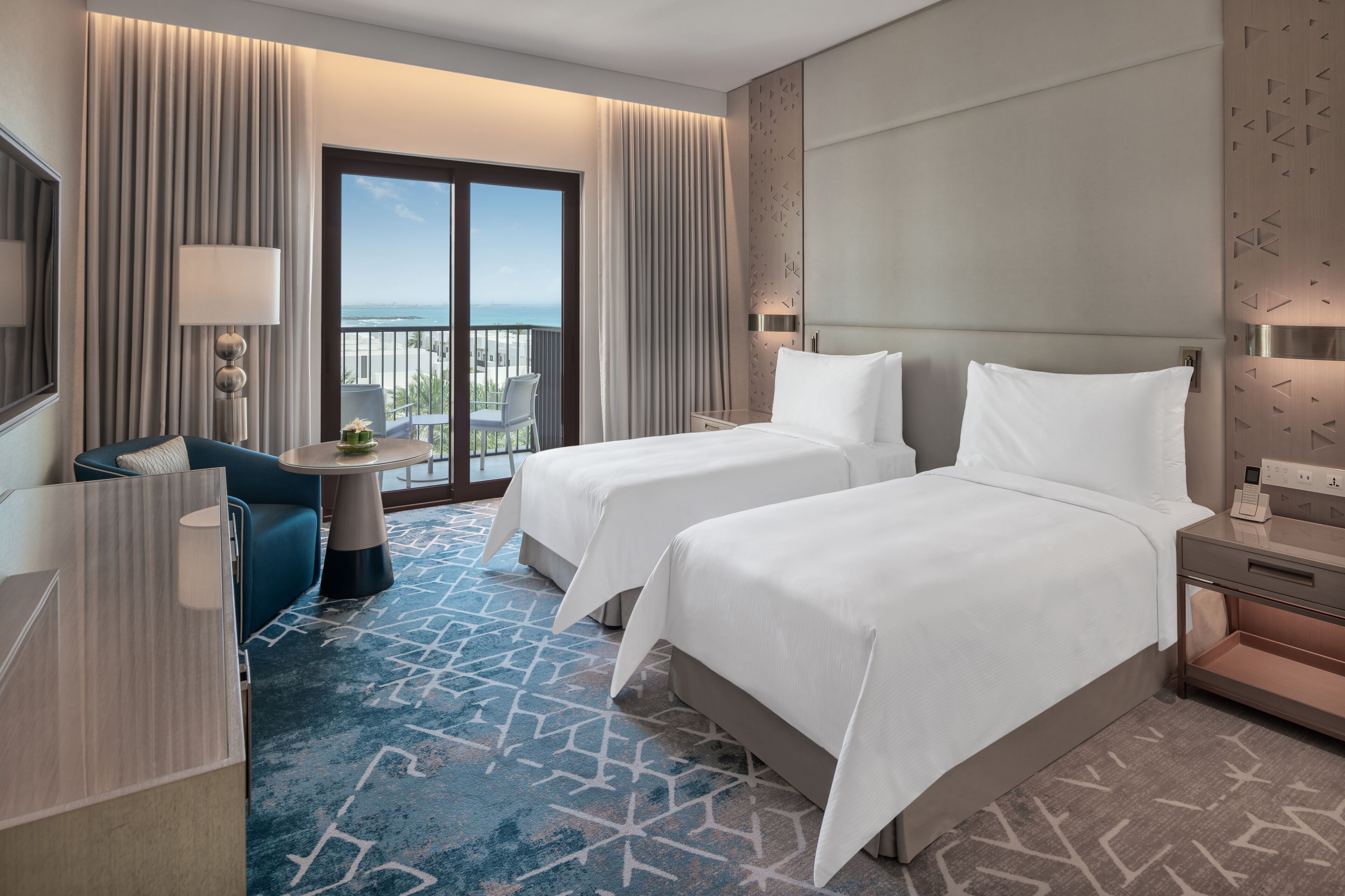 A hotel room with two beds, a desk, a chair, a balcony with a view of the sea, and a patterned carpet.