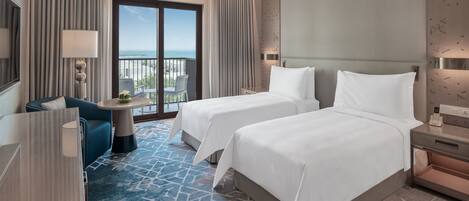 A hotel room with two beds, a desk, a chair, a balcony with a view of the sea, and a patterned carpet.