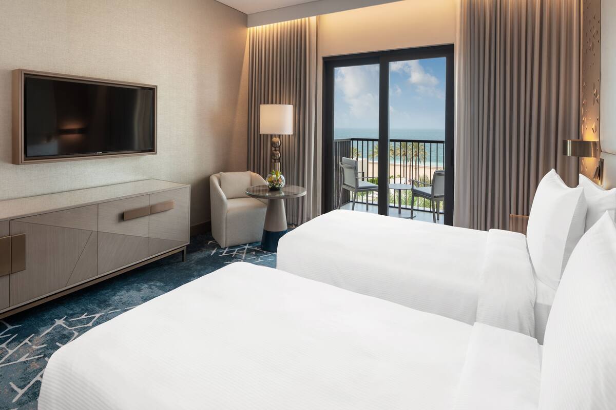 Une chambre d’hôtel avec un grand lit, un téléviseur fixé au mur, une petite table avec une lampe et un balcon donnant sur l’océan.