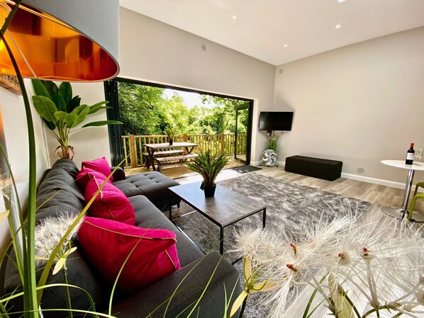 A modern living room with a sofa, coffee table, and a dining area with a table and chairs. There is a TV mounted on the wall and a potted plant on the coffee table.