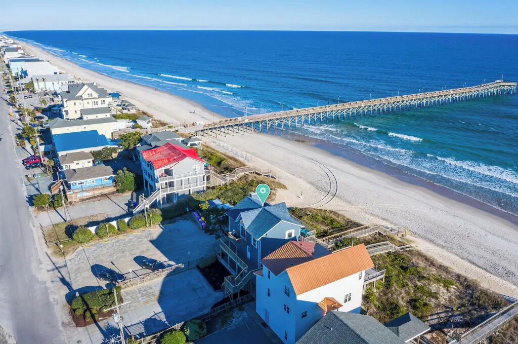 An aerial view of a coastal town with a pier extending into the ocean, beachfront properties, and a clear sky.