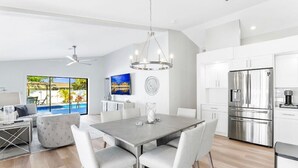 A modern kitchen with stainless steel appliances, a dining area with a grey table and white chairs, and a large window offering a view of a swimming pool. - Luxurious Modern 4BR, Heated Pool near Beach and Mercato! Newly Renovated! (Naples)