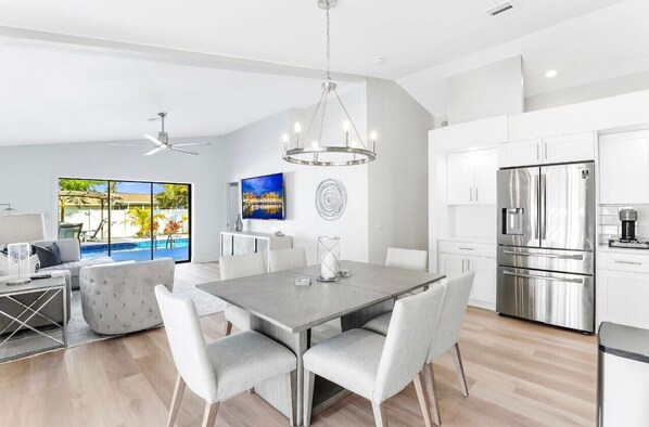 A modern kitchen with stainless steel appliances, a dining area with a grey table and white chairs, and a large window offering a view of a swimming pool.
