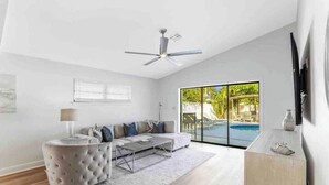 A modern living room with a sofa, coffee table, and ottoman, a ceiling fan, and a large sliding glass door leading to a pool area. - Luxurious Modern 4BR, Heated Pool near Beach and Mercato! Newly Renovated! (Naples)