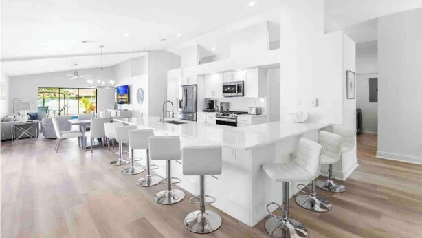 A modern kitchen with white cabinetry, a central island with bar stools, and a dining area with a gray sofa and white chairs. - Luxurious Modern 4BR, Heated Pool near Beach and Mercato! Newly Renovated! (Naples)
