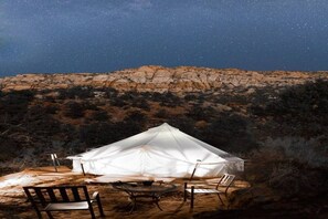 Une tente blanche, avec une table et des chaises installées, se dresse au milieu d’un paysage désertique, de nuit.