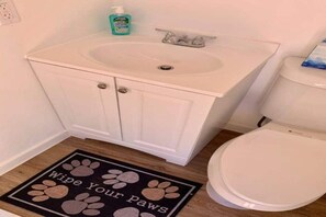 Une salle de bain avec un lavabo blanc, des toilettes et un paillasson orné d’empreintes de pattes et de l’inscription « Essuyez vos pattes ».