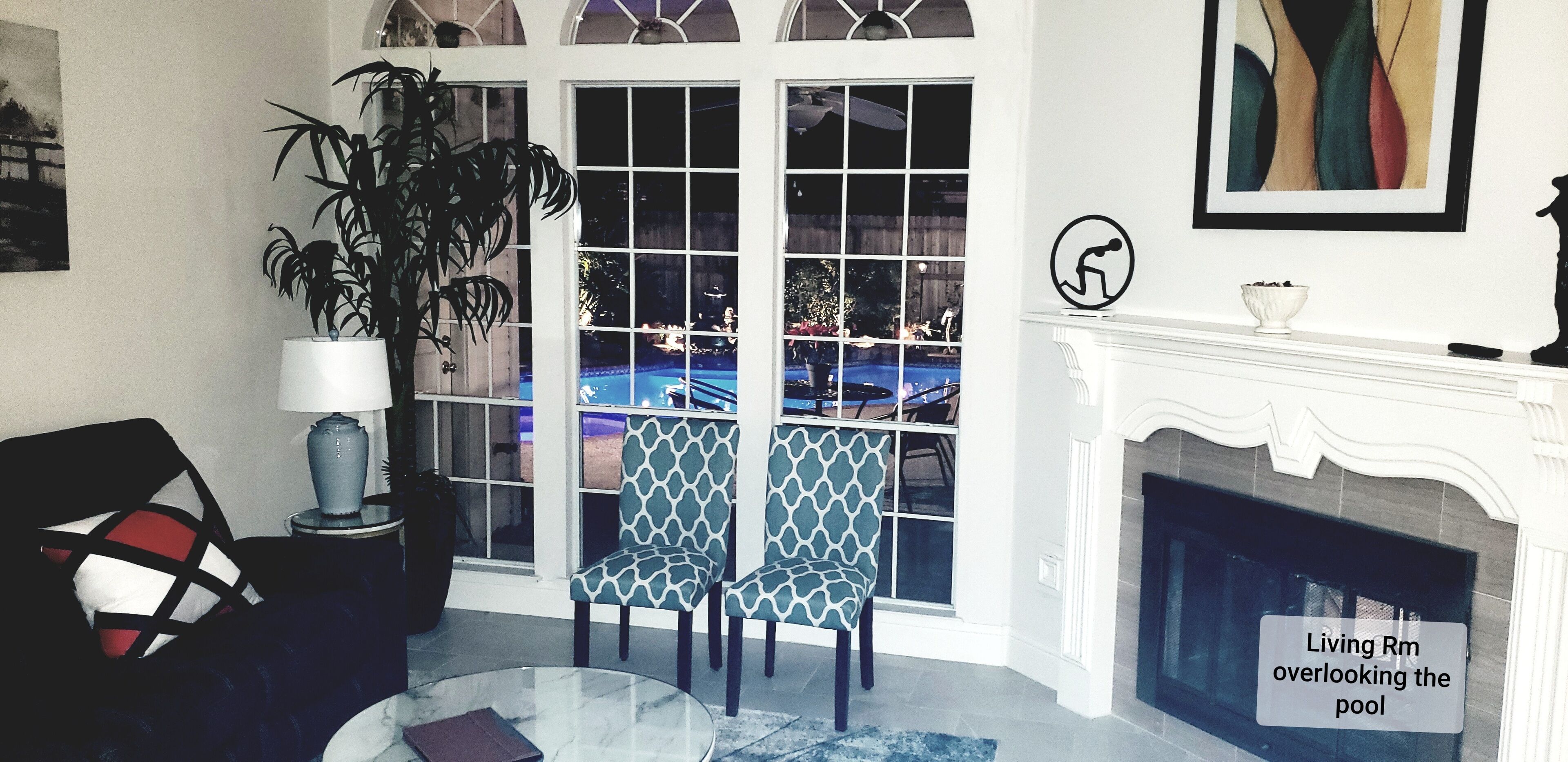 A living room with a glass coffee table, two patterned chairs, a sofa, a lamp, a fireplace, and a view of a pool through large windows.