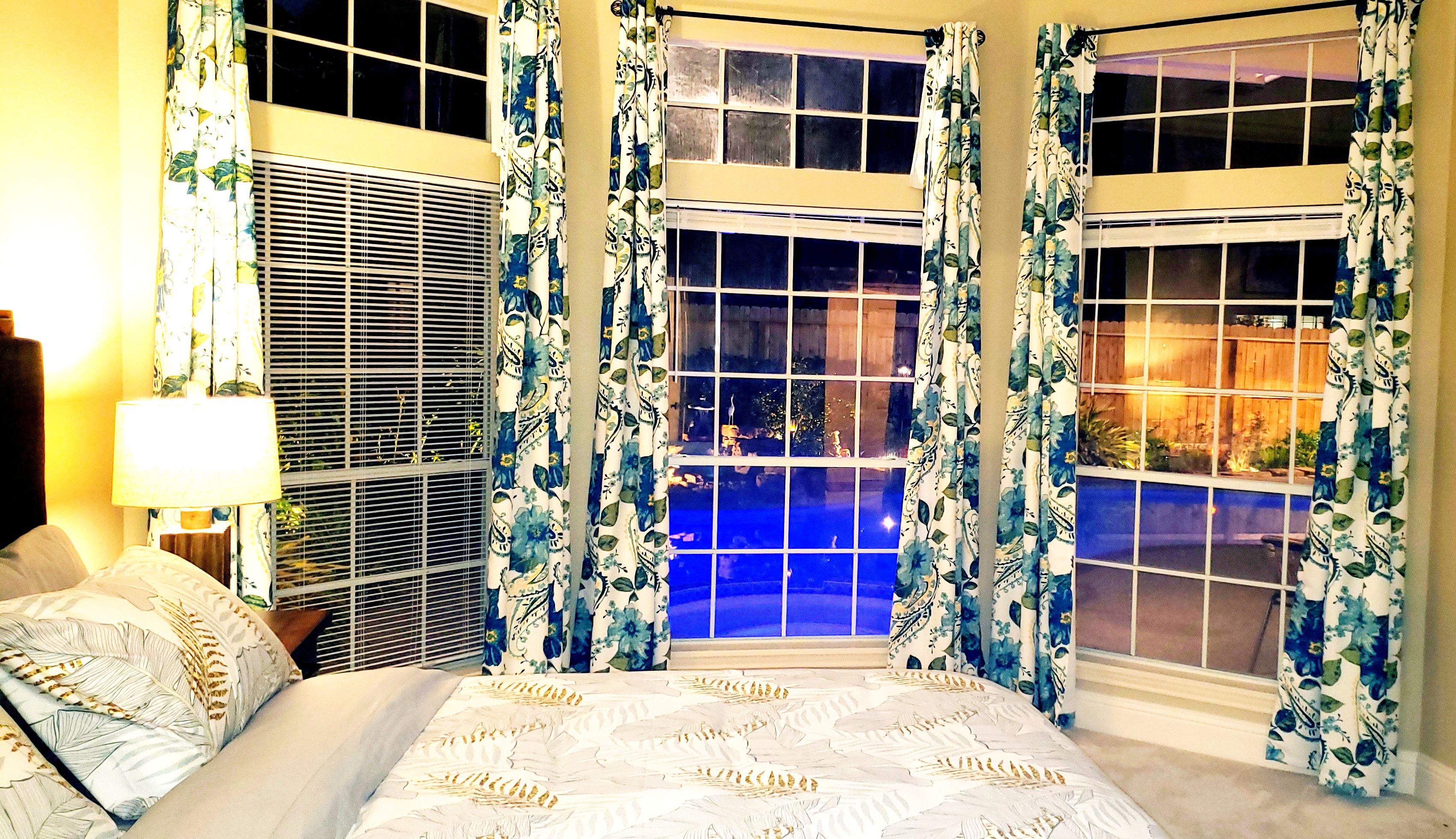 Un dormitorio principal con una cama grande, una mesita de noche con lámpara y vistas a una piscina a través de las ventanas.