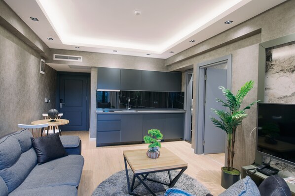 A modern living room with a sofa, coffee table, and a TV. There is a kitchen area with cabinets and a sink. - Marlen Residence Hotel (Izmir)