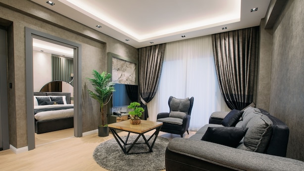A modern living room with a sofa, armchair, and a small wooden coffee table. There is a bed in the background and a potted plant on the coffee table. - Marlen Residence Hotel (Izmir)