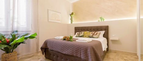 Un dormitorio con cama, una planta en maceta, un tapete colorido y una lámpara de pared.